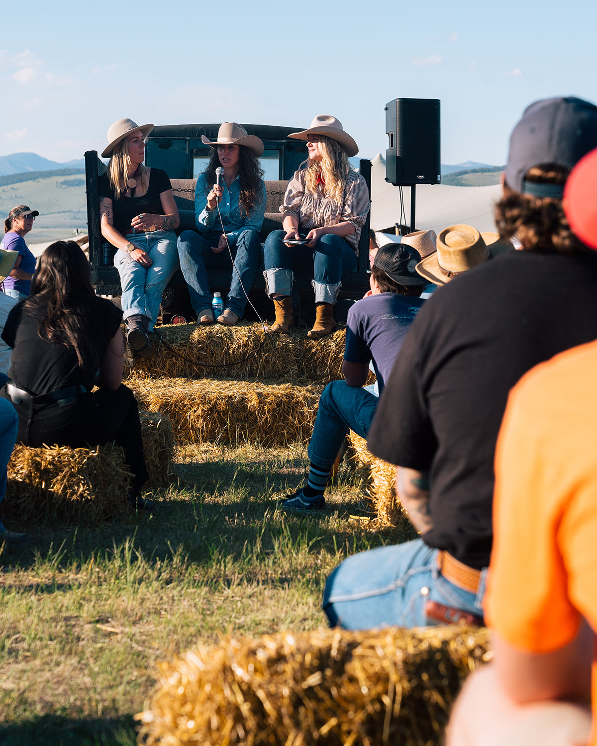 Three women in cowboy hats speaking on a hay-bale stage to an outdoor audience